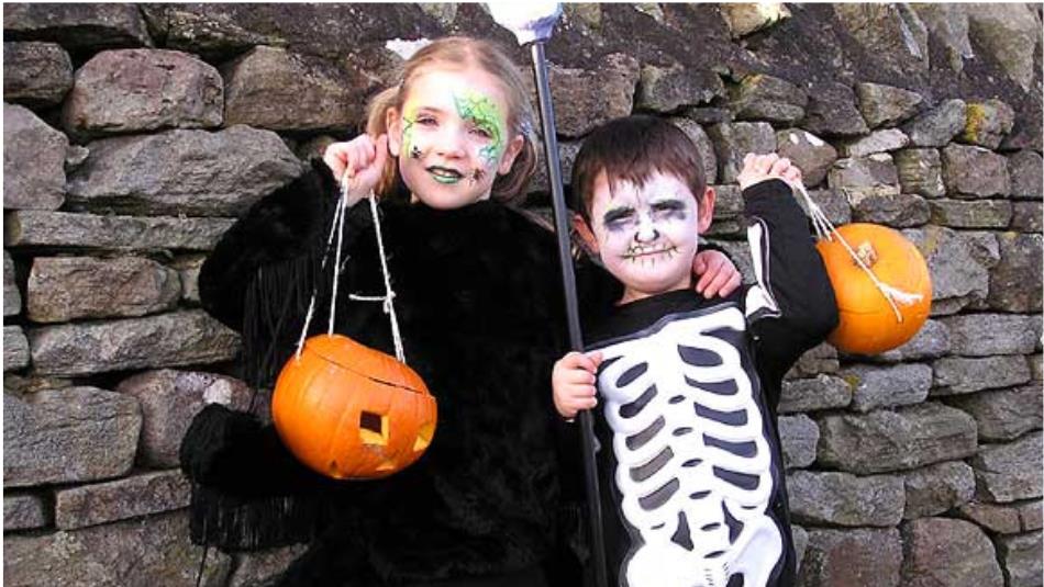 Two children dressed up as a witch and a skeleton, smiling holding pumpkins against a stone wall.