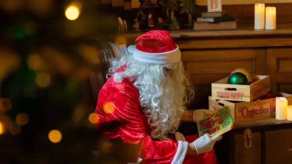 Santa Claus sitting near a Christmas tree, reading a book with toys.