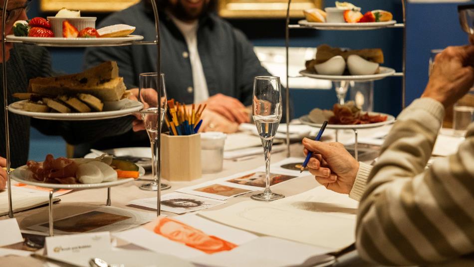 Photograph captures a table table during a shared meal, with small plates and diners eating, drawing and selecting food from tiered serving stands. Pl