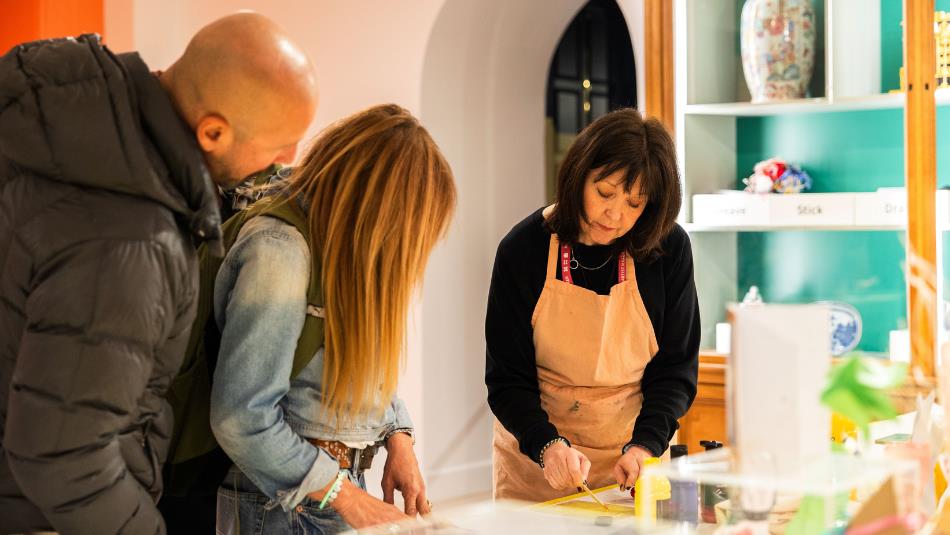 Photograph shows an art workshop or demonstration, with two visitors observing a woman engaged in an activity at a table. The woman doing the demonstr