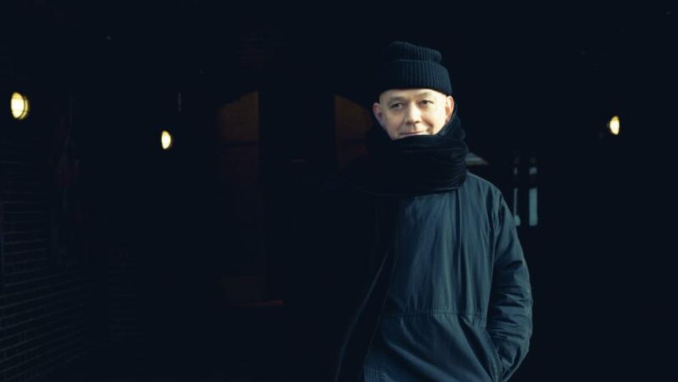 A person wearing dark clothes, a hat and scarf, standing smiling in a dark hallway.