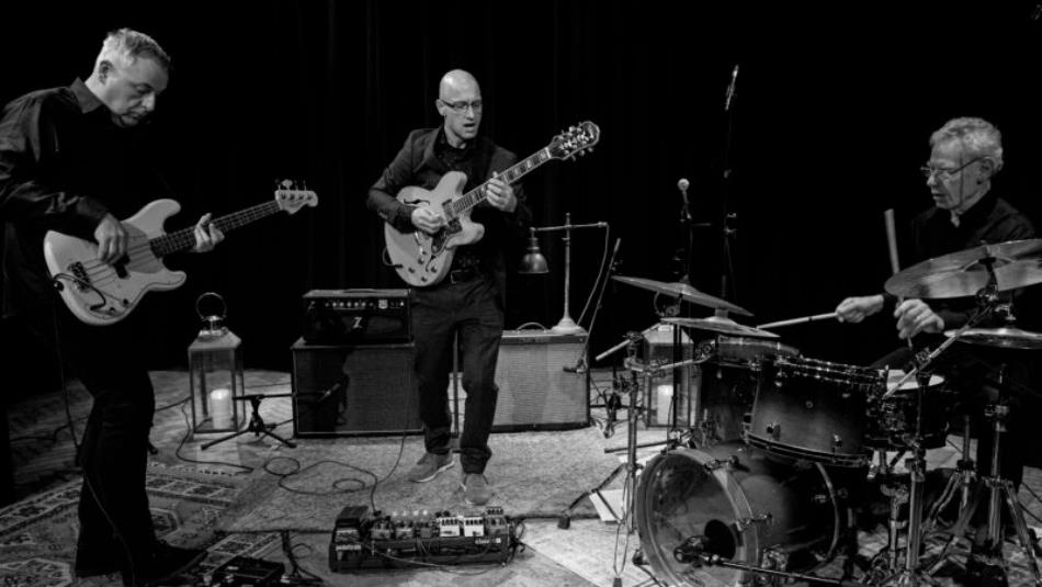 Three people playing their respective instruments, bass, guitar and drums in a rehearsal space. The image is in black and white.