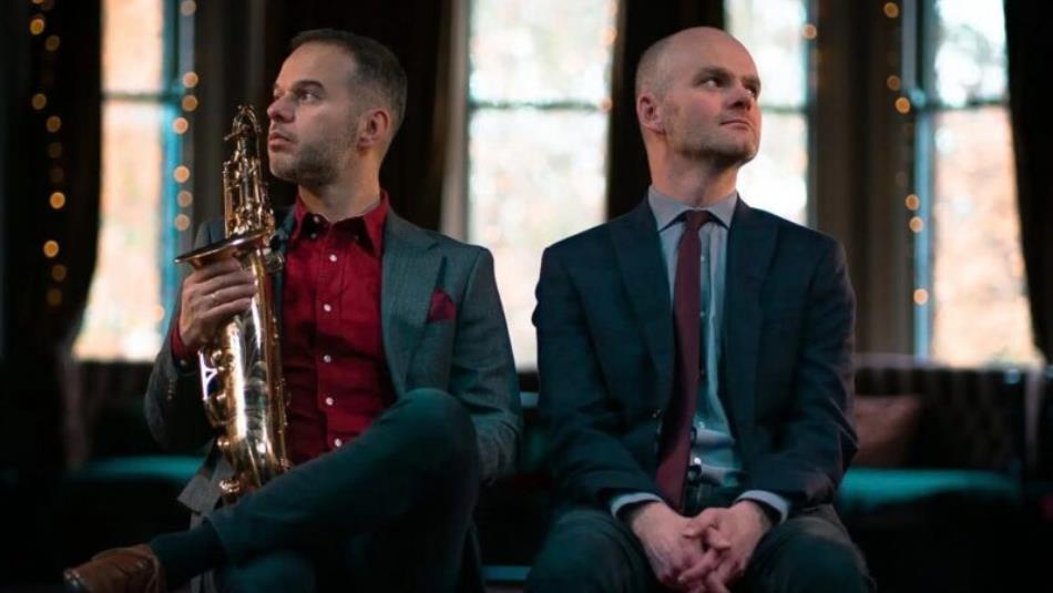 Two people sat facing away from each other, wearing suits and holding a brass instrument.
