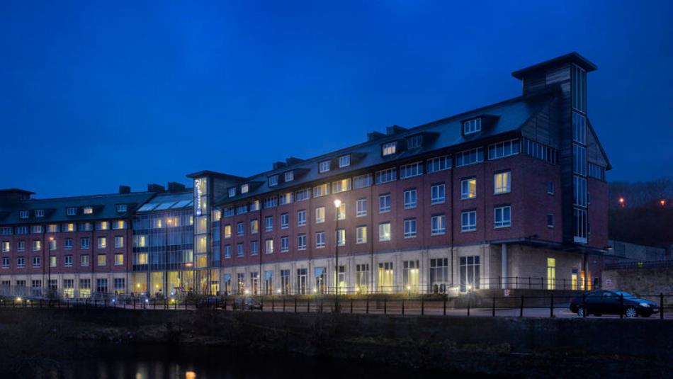 A large hotel building on the side of a river, in the dark with glowing windows.