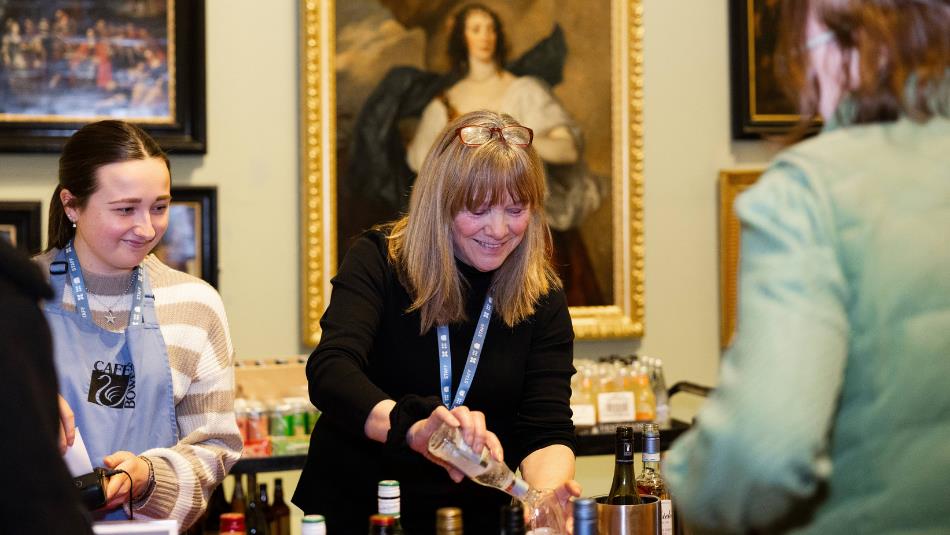 Photograph of women conducting a food or drink tasting at a museum event, with bottles, samples and name badges arranged on a table. A large framed po