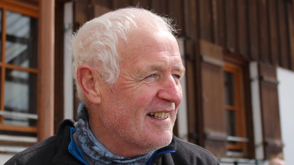 Photograph showing an older white-haired man outdoors beside a timber-clad building. He smiles in profile, wearing a black jacket and blue-grey neck w