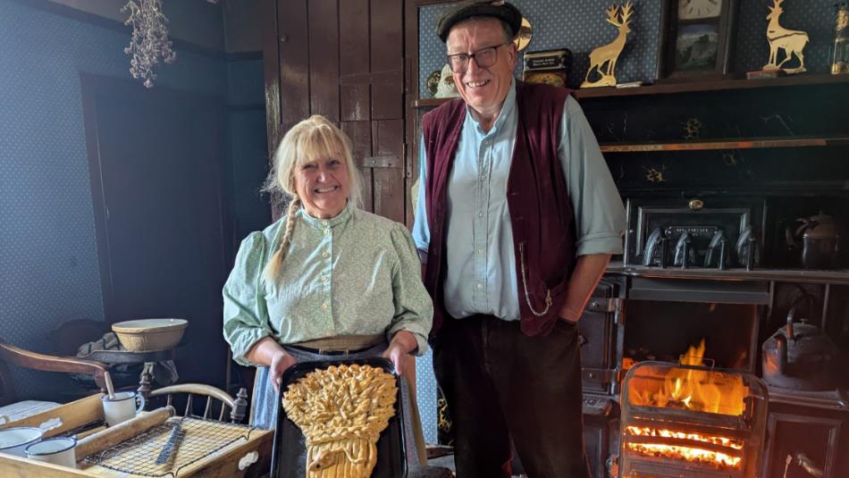 Two people standing in the kitchen of a historic house, holding a harvest style baked loaf.
