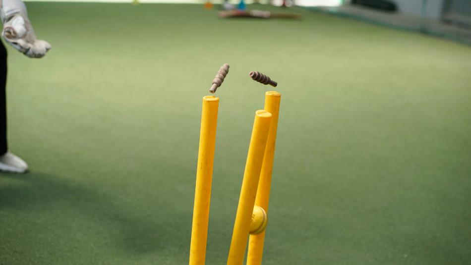 Yellow cricket stumps being hit by a yellow cricket ball, on a green lawn.