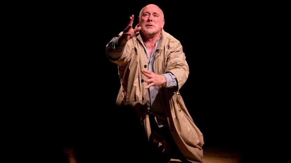 A person wearing a beige trench coat kneels on a dark stage looking up in fear.