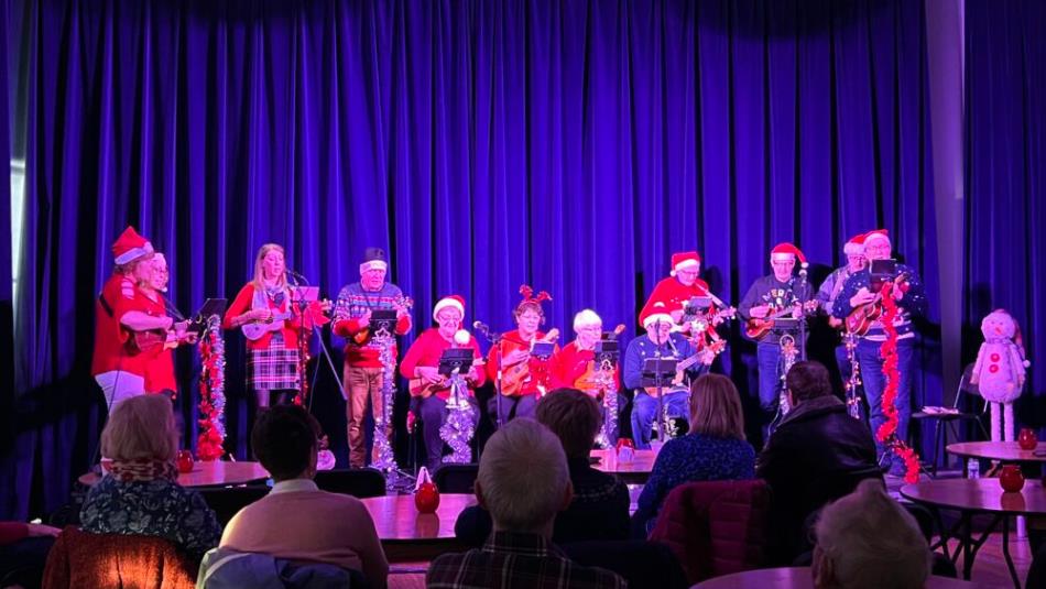 A group of festive performers wearing Santa hats and Christmas jumpers playing the Ukulele on a blue stage.
