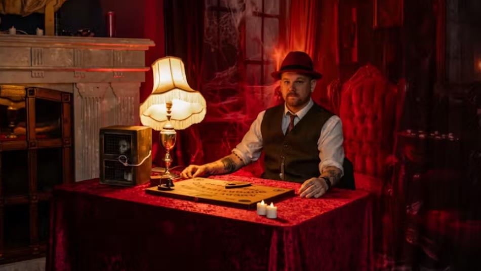 A person in a hat and waist coat, sat at a vintage-style desk with a Ouija board, surrounded by red sinister mist.