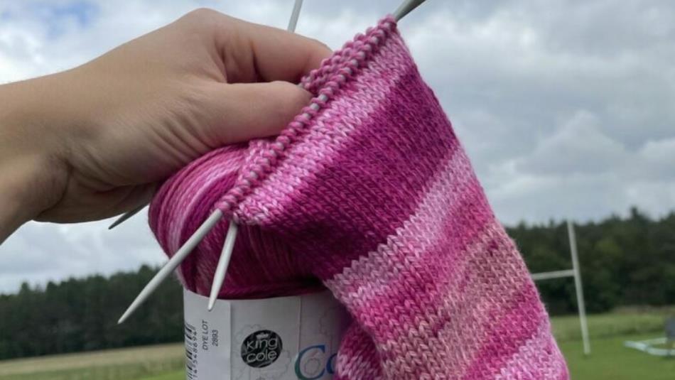 A pink knitted piece and ball of wool held up outside.