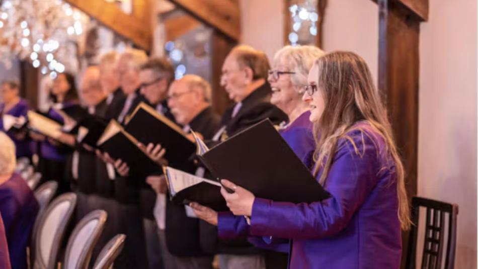 A group of festive choir singers, wearing purple and holding song books.