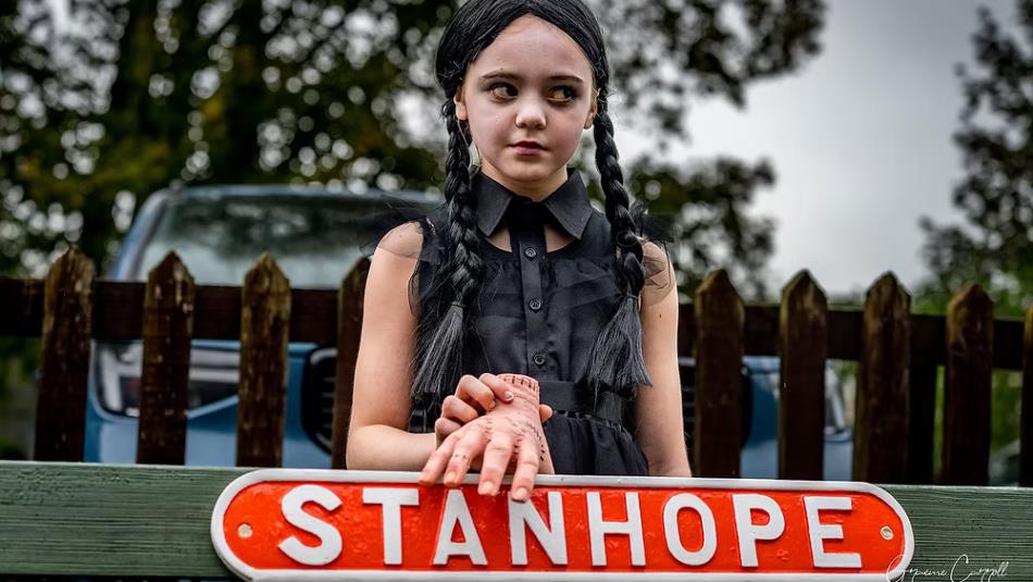 A Wednesday Addams character standing in front of a Stanhope railway sign with a fake severed hand.