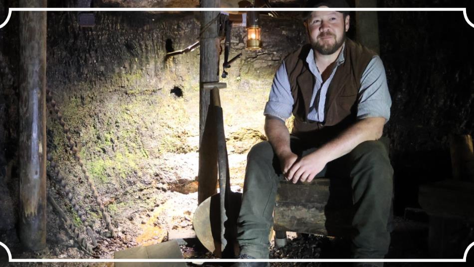 A person wearing historical mining clothes sat in a dark pit, smiling, surrounded with tools.