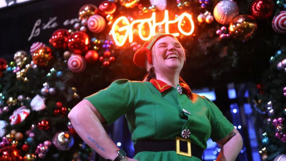A person dressed as a festive elf, looking up and smiling in front of a lit up sign which reads, 'Grotto' decorated with baubles.