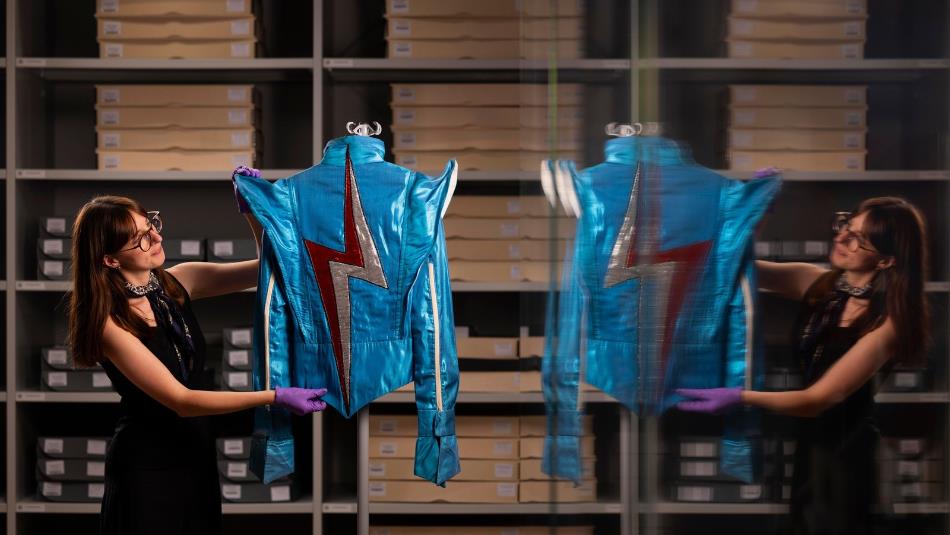 Photograph showing a curator in purple gloves displaying a bright blue metallic costume inside a clothing archive. Shelves of labelled storage boxes a
