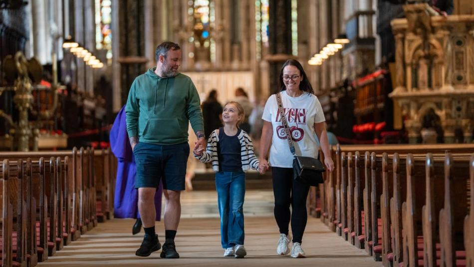 Two adults walk either side of a child holding hands down the pews of Durham Cathedral.