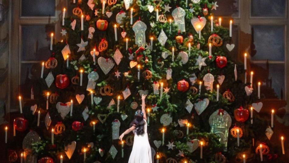 A scene from The Nutcracker ballet of a dancer looking up, with their back to the stage with their arm elegantly raised at a large christmas tree.
