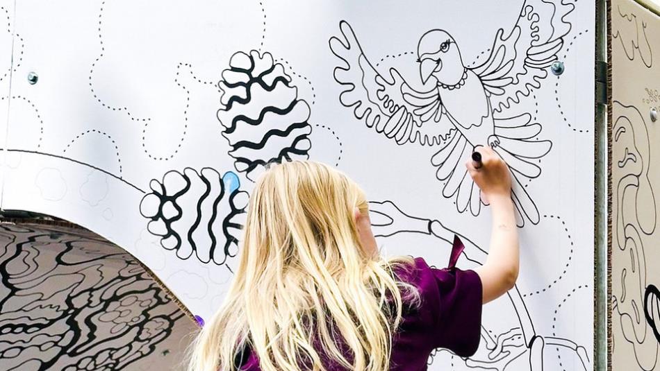 Photograph shows a child in a purple top drawing on a illustrated wall while sitting on an adult's shoulders. Wall features black line-art birds, fish