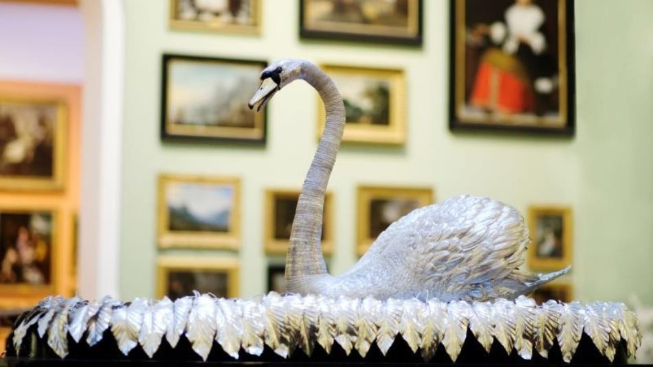 Photograph of a silver swan displayed in a gallery or museum, surrounded by framed artworks. Curved neck, textured feathers and ornate leafy base form