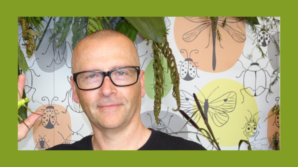 A person smiling surrounded by plants, holding a stick insect on their hand, on a green background.