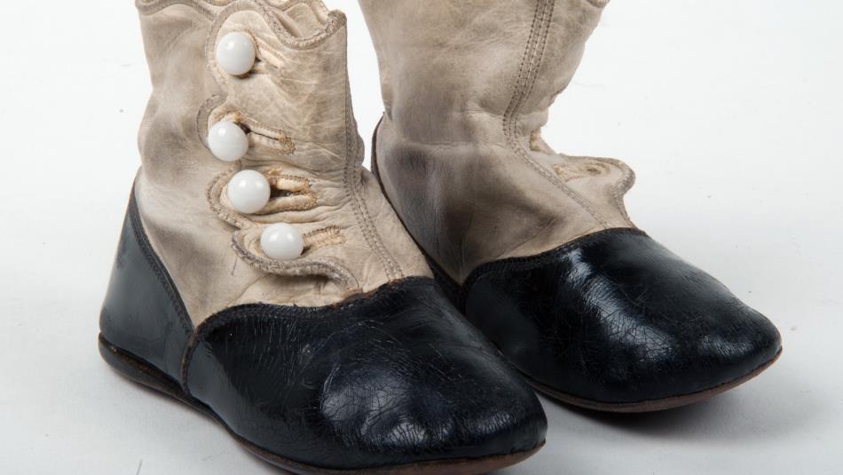 A pair of historic children's ankle boots is displayed against a plain white background. The boots are made from cream-coloured leather and black mate