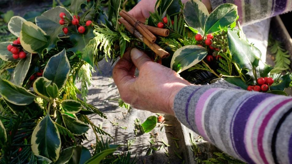 A pair of hands tending to a festive wreath, full of holly, red berries and cinnamon sticks.