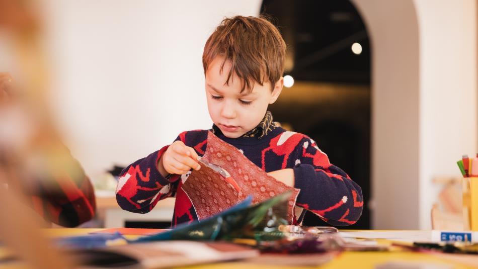 Photograph of young child creating a craft project at a table, using scissors and patterned red paper. Colourful materials, brushes and tools surround