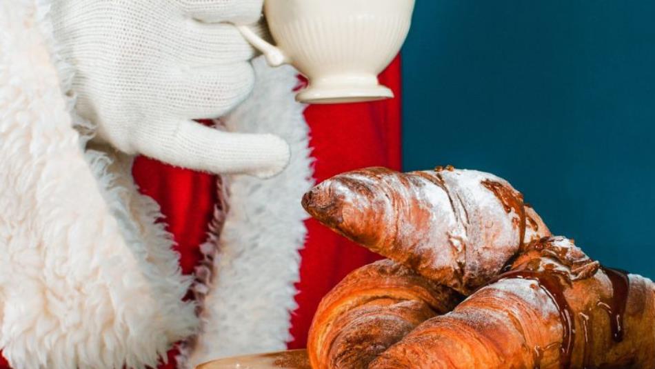 Close up view of Santa's hand holding a teacup with his little finger pointing out, in front of a plate of croissants.