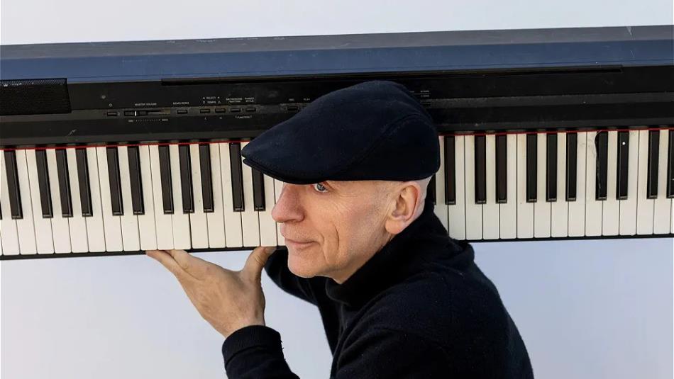 Person wearing a black flat cap and a turtle neck, carrying a piano keyboard over their shoulder against a grey background