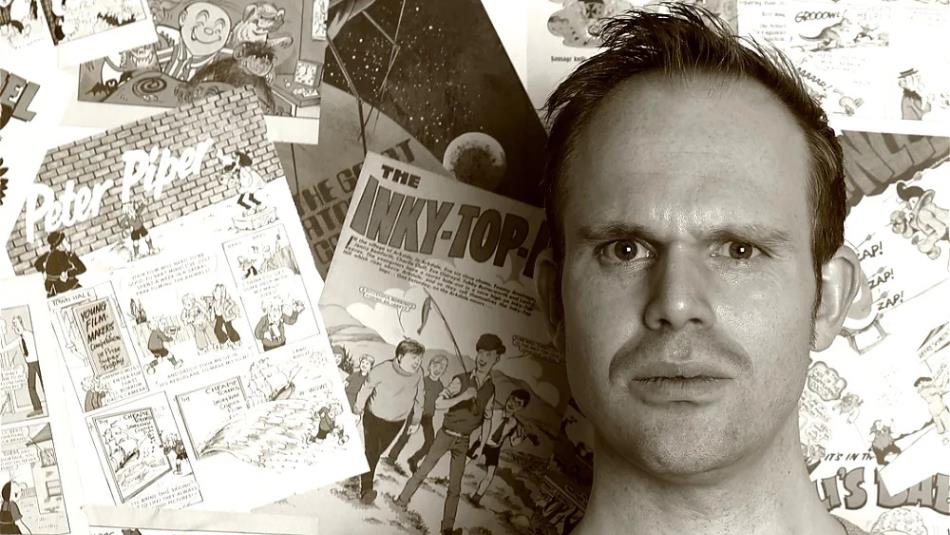 Alex Collier looking quizzical, shoulders up, in front of a wall of vintage comic books, the image is in black and white.