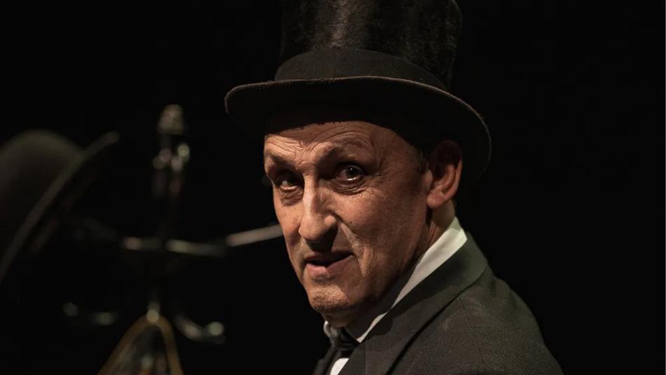 Man wearing a top hat and suit, looking over his shoulder with a shocked expression, shrouded in darkness.