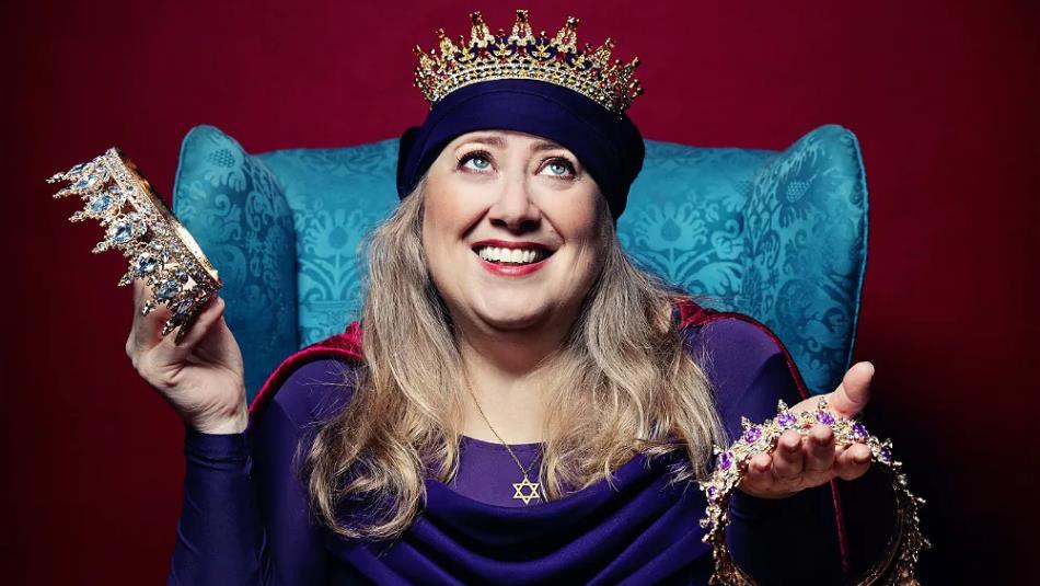 Person wearing a crown, sat on a blue chair holding two crown other crowns on a burgundy background.