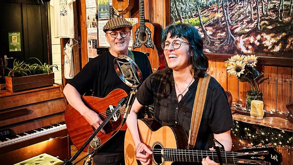 Two people playing guitars, smiling in a folk-like venue