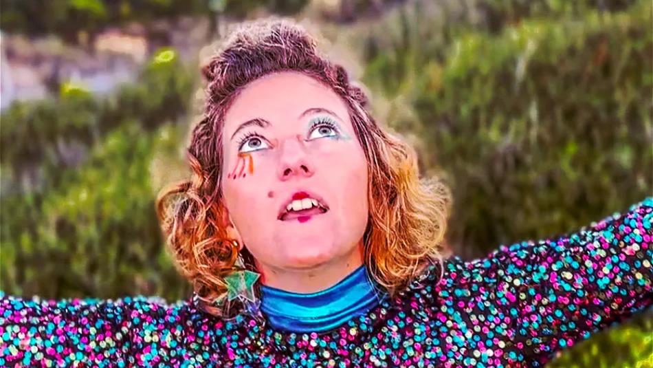 Person wearing colourful sequins and makeup, looking up bewildered with arms open, in front of nature.