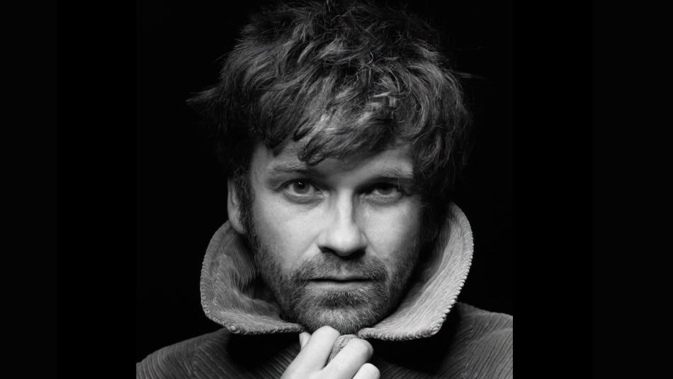 Black and white photograph of a man with tousled hair and a beard, looking directly at the camera.