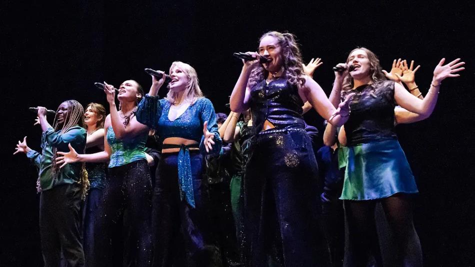 Group of people on a dark stage, singing wearing sparkly blue outfits