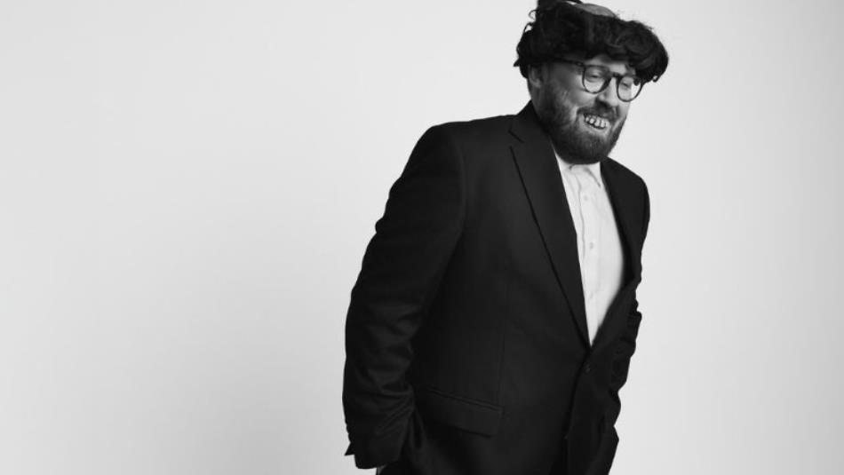 Comedian John Kearns stood leaning to one side wearing joke teeth and a wig. The image is in black and white