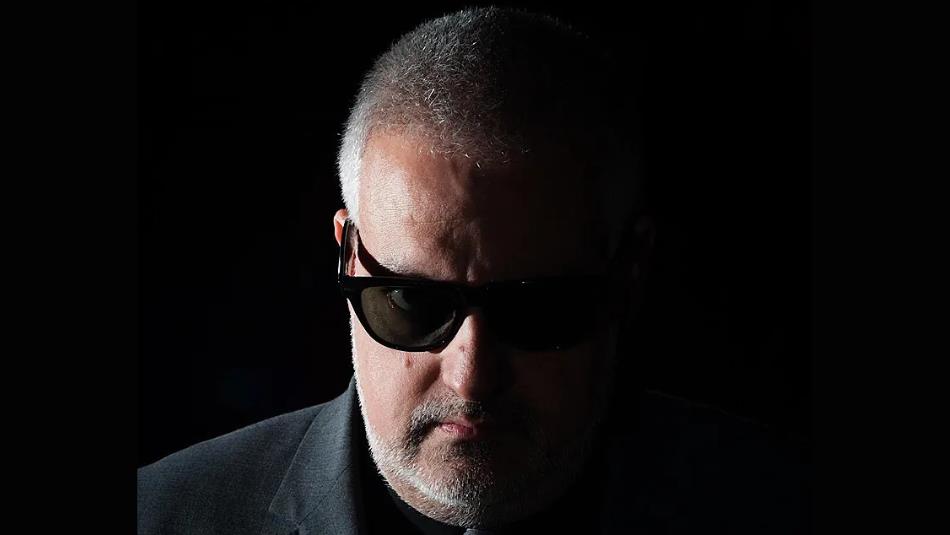 A person shrouded in darkness, looking sternly at the camera wearing sunglasses