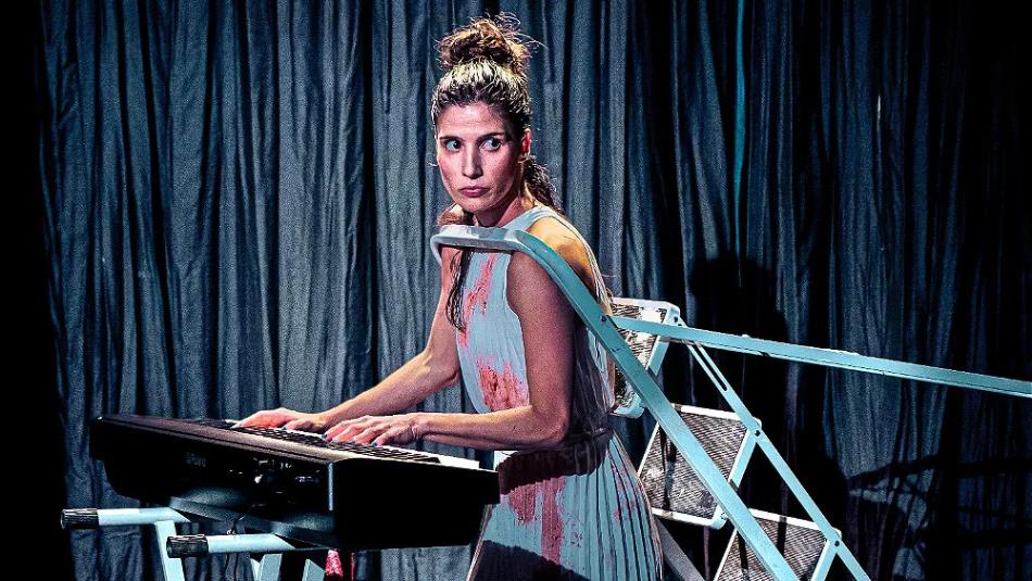 A person stood playing the keyboard, stood in front of a blue curtain with a step ladder around their shoulders
