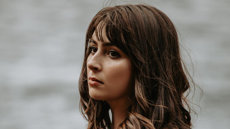 Photograph of a woman with shoulder-length brown hair, wearing a black top, looking pensively to her left. Background features a softly blurred body o