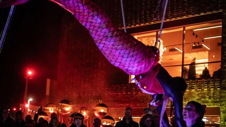 Person suspended on wires wearing a pink mermaid tail holding hands with someone
