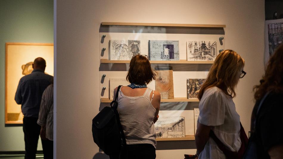 People observing framed sketches and drawings displayed on a gallery wall, highlighting an art exhibition setting.