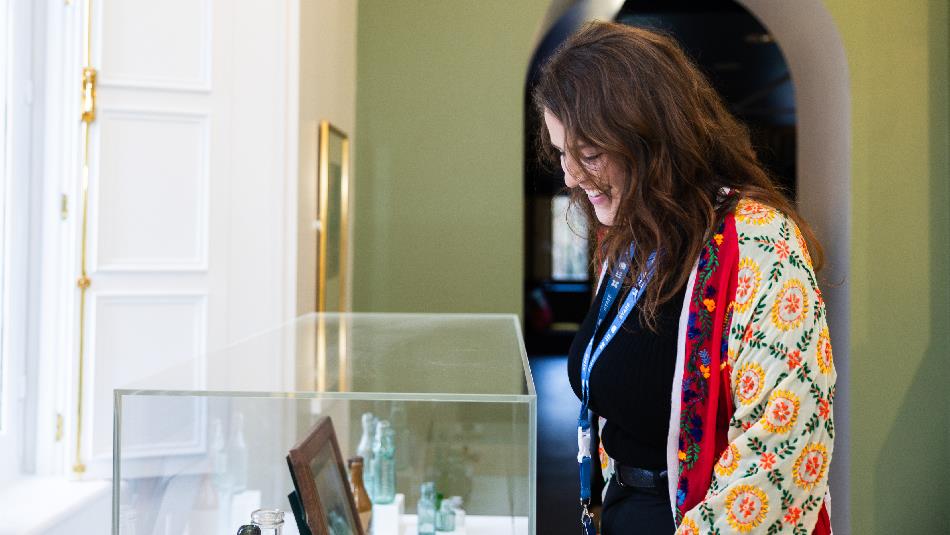 A woman wearing a colourful embroidered jacket smiles as she looks into a glass display case filled with vintage bottles and framed items in a museum