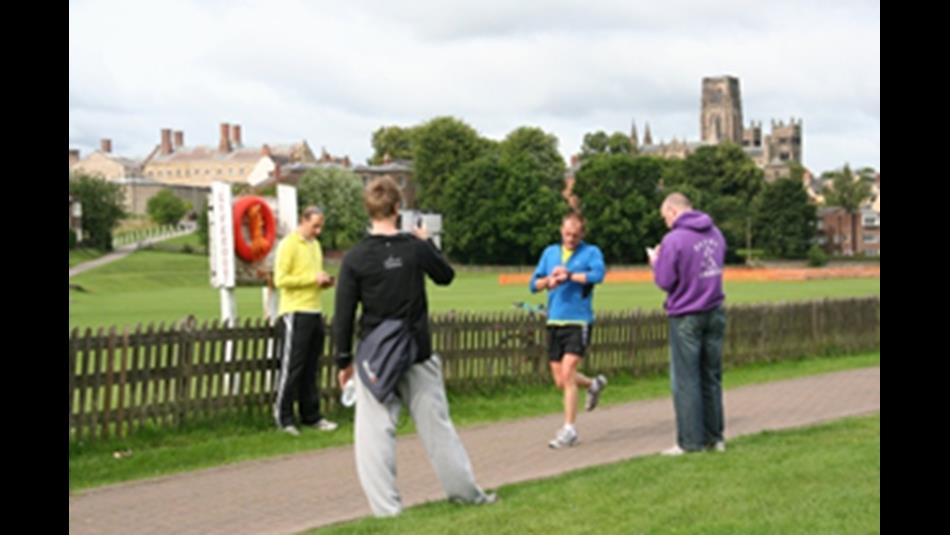 Runners partaking in Park Run by the river, with Durham Cathedral in the background.