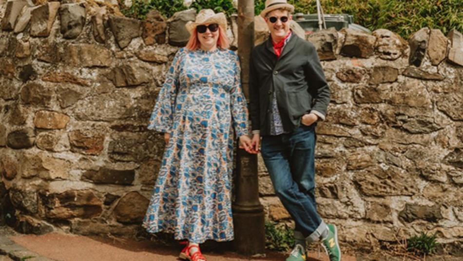 Founders of For the Love of the North Paul and Lucy Hull stand hand-in-hand beside an old stone wall and a vintage signpost in a quaint village.