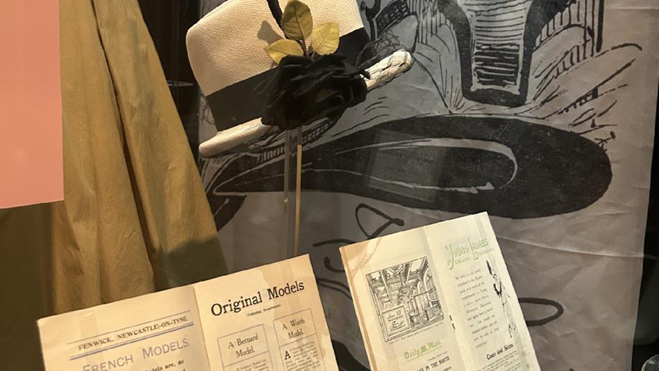 A cream hat with black ribbon on a stand alongside archival Fenwick material