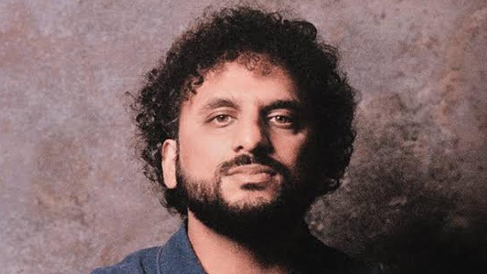 Comedian, Nish Kumar, stands straight faced in front of a purple shadowy background.