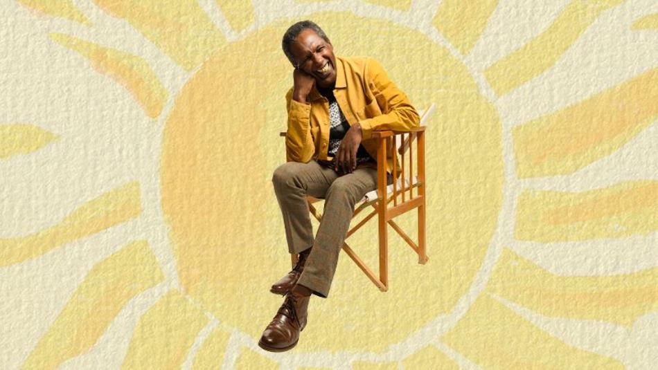 Lemn Sissay sits on a wooden chair, smiling, cut out in front of a painted yellow sun.